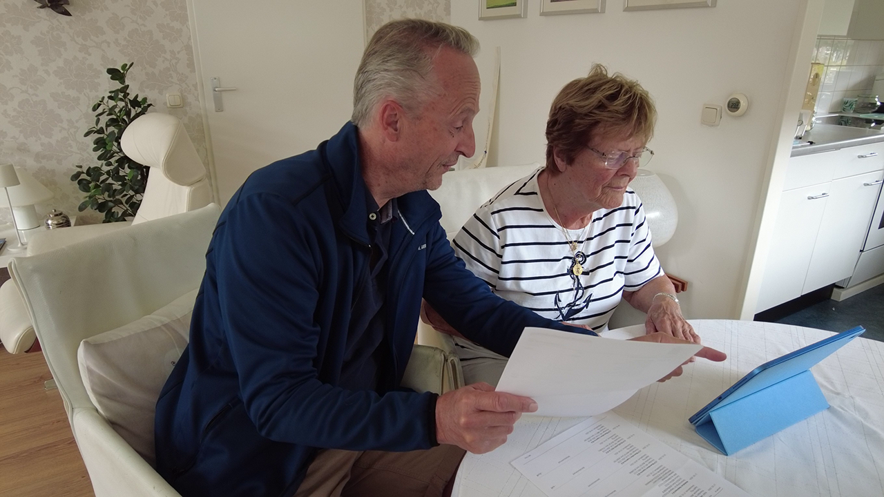Oudere man helpt een oudere vrouw aan de eettafel bij het bekijken van documenten en een tablet in een rustige thuisomgeving.