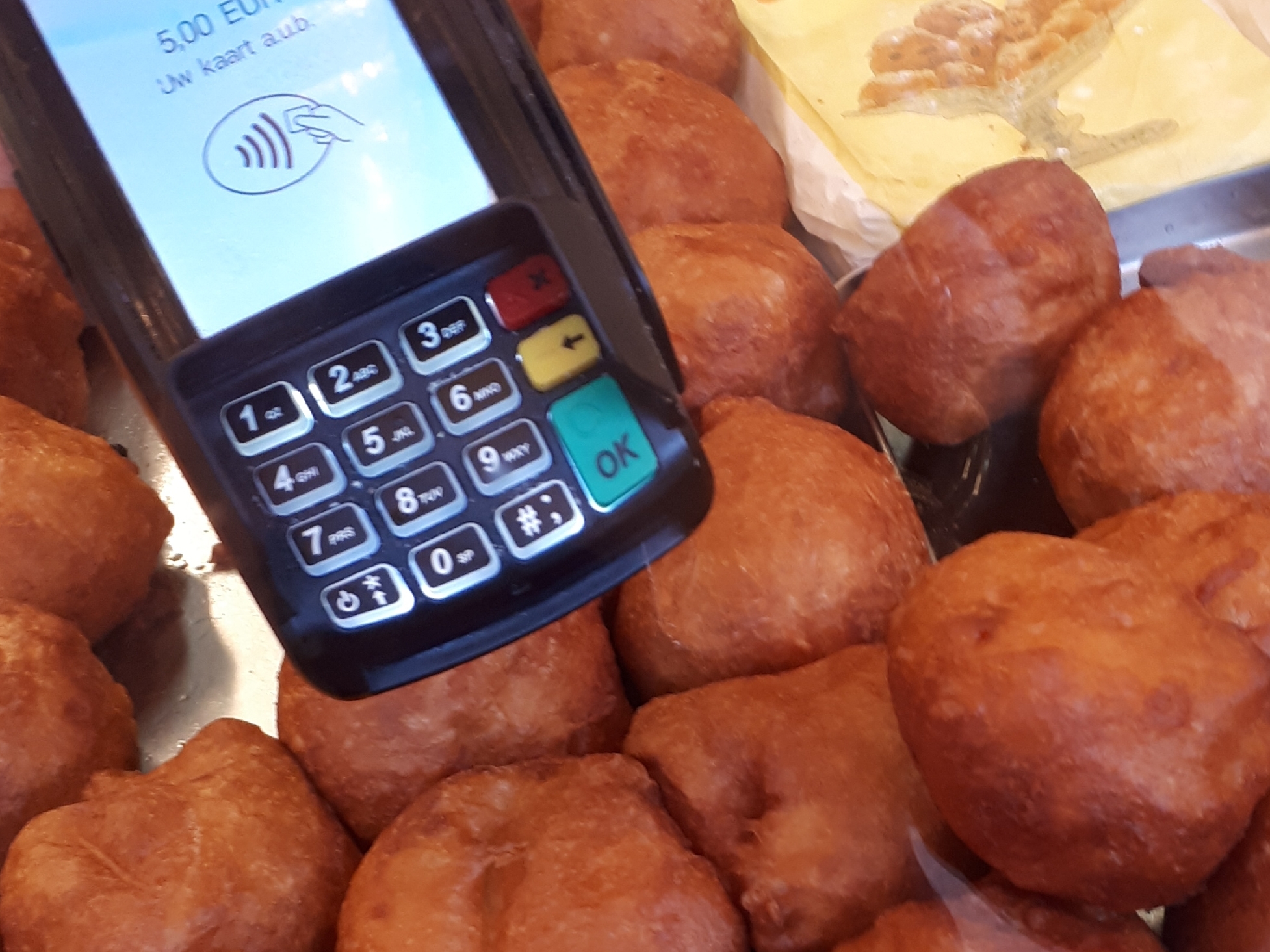Close-up van pinterminal boven schaal vol oliebollen; scherm toont bedrag van 5 euro en symbool voor contactloos betalen.