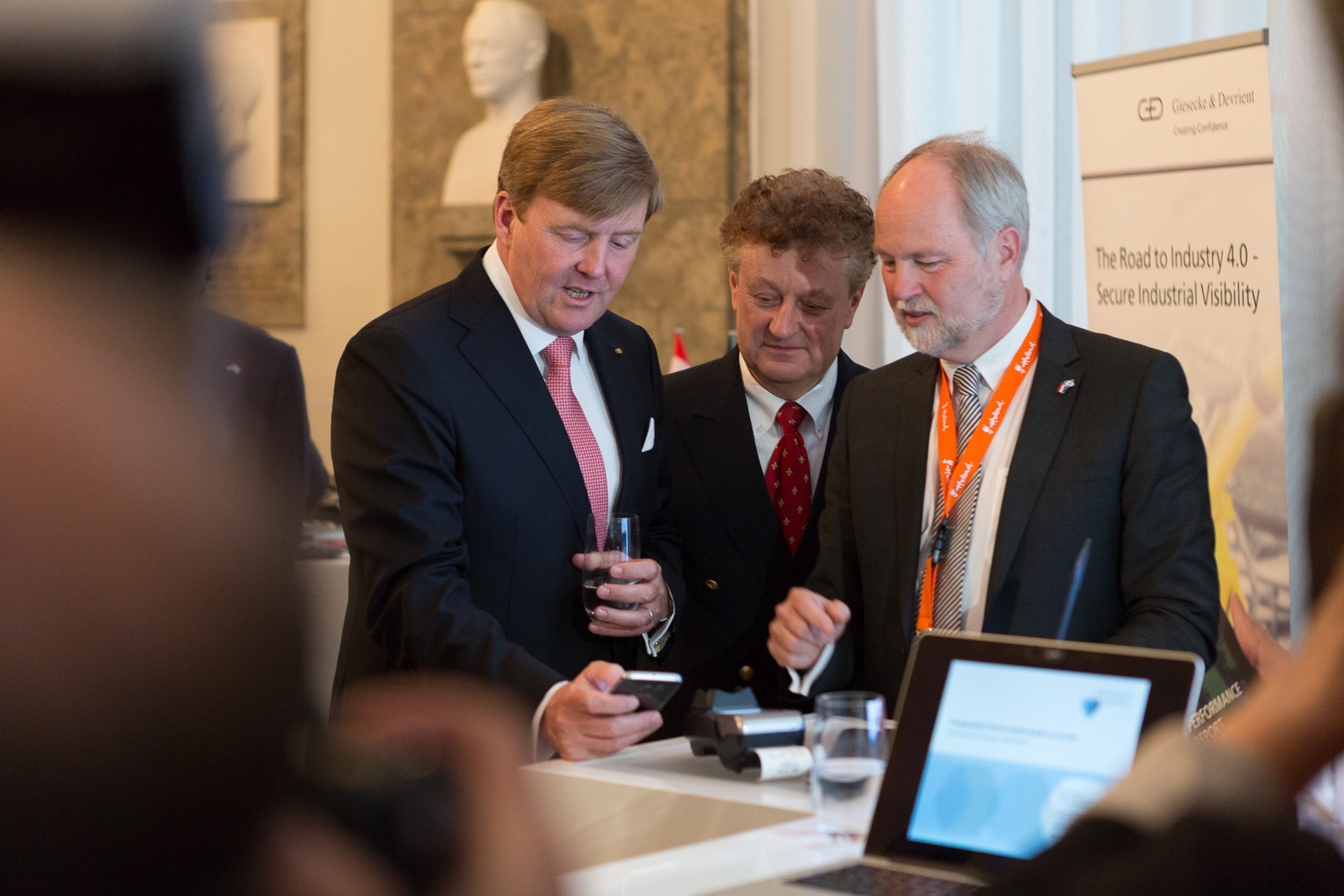 Koning Willem Alexander bespreekt iets op een smartphone bij statafel met laptop tijdens een zakelijke bijeenkomst.