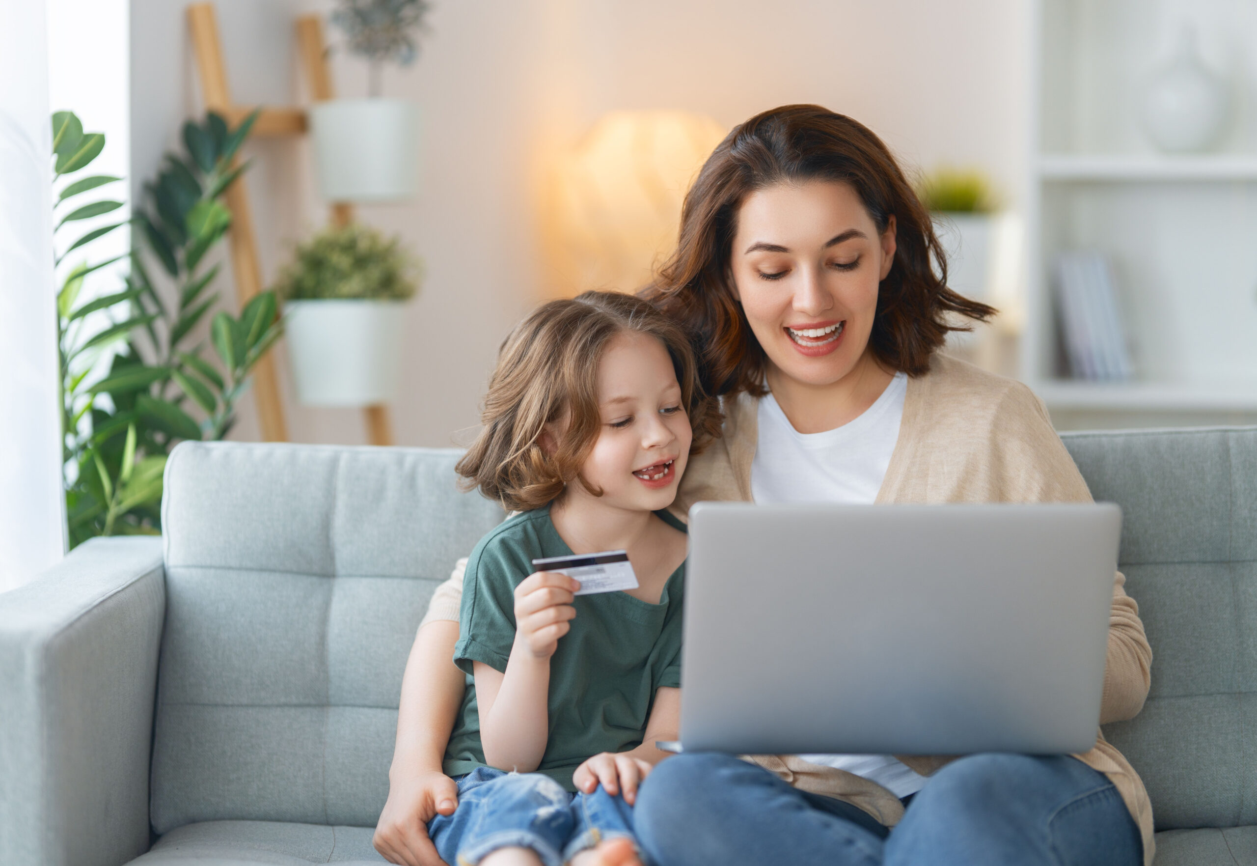 Moeder en jong kind zitten samen op de bank en doen een online betaling op een laptop, terwijl het kind een betaalkaart vasthoudt in een huiselijke sfeer.