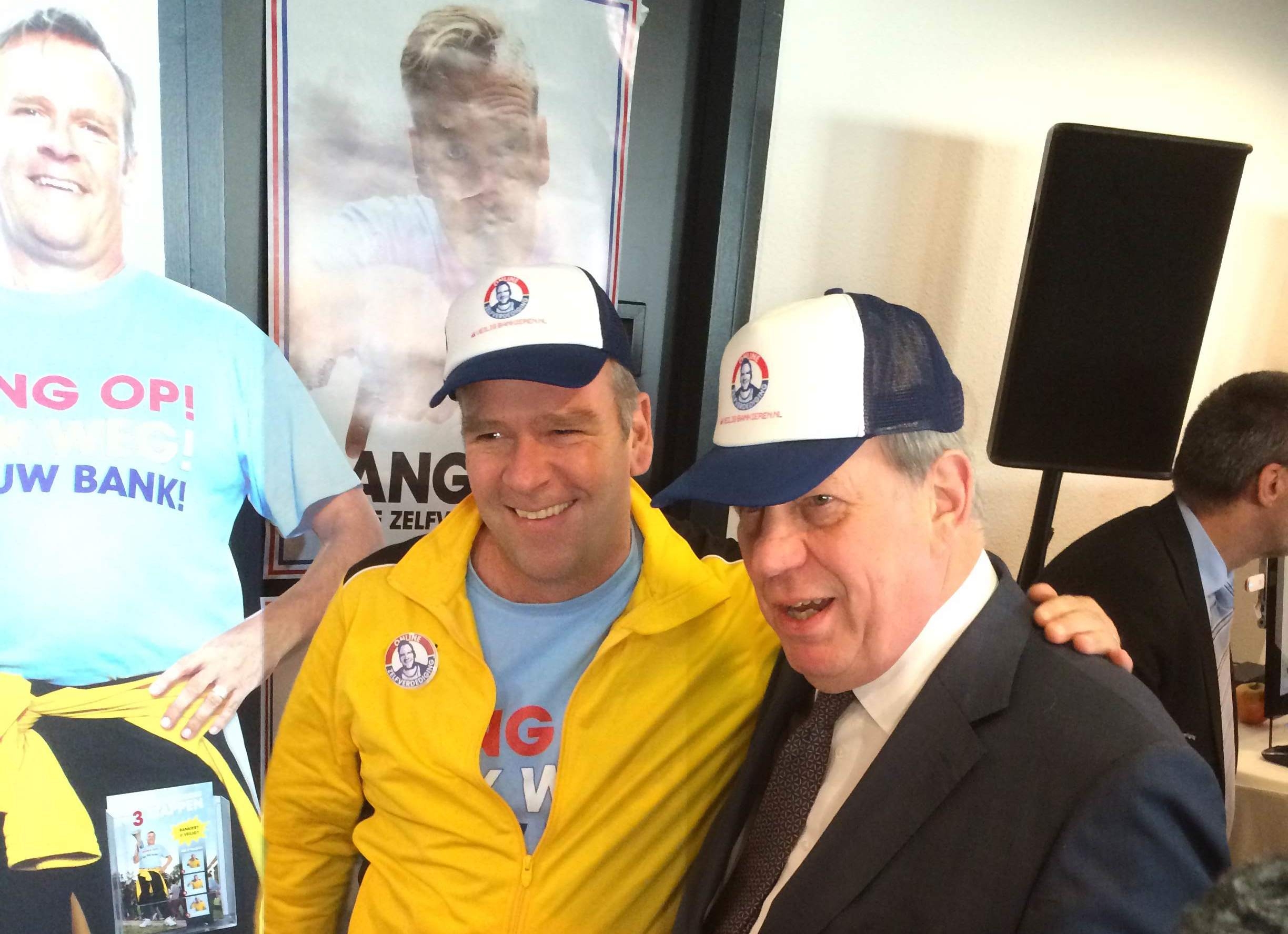 Twee mannen met campagnepetten poseren lachend voor een promotiewand met slogan over veilig bankieren in een drukke zaal.