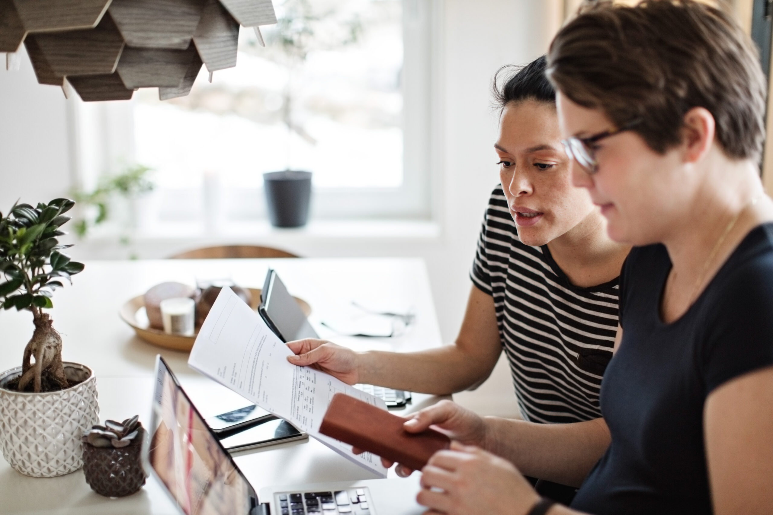 Twee collega’s bespreken een rekening aan tafel, vergelijken papieren gegevens met laptop en smartphone in een thuiswerkplek.