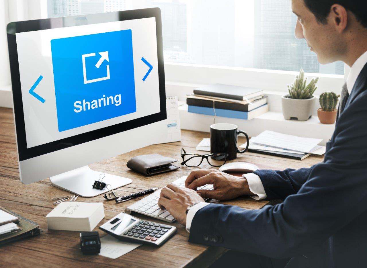 Zakelijke man werkt aan bureau op kantoor, terwijl groot computerscherm met blauwe tegel ‘Sharing’ veilig datadelen benadrukt.