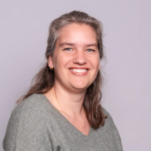 Glimlachende vrouw met lang bruin haar in een grijze trui, gefotografeerd tegen een lichtgrijze achtergrond.